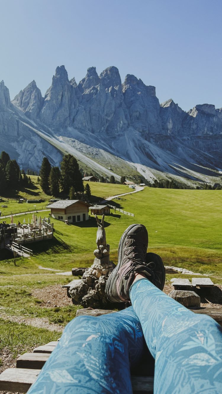 chica tumbada que solo se ven piernas con botas de montañas hacia un mirador de prado verde y de cadena montañas al fondo Refugio Odle